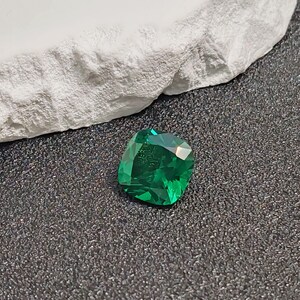 May include: A vibrant, emerald-green gemstone with a cushion cut. The gem is displayed on a dark, textured surface, with a piece of white, rough-textured material in the background. The gem's facets reflect light, showcasing its clarity and color.