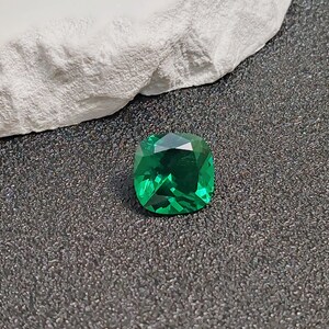 May include: A vibrant, emerald-cut green gemstone with a faceted surface. The gem is displayed on a dark, textured surface, with a white, rough-textured element in the background. The gemstone's facets reflect light, creating a sparkling effect.