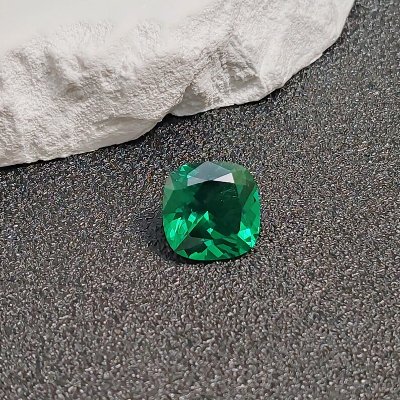 May include: A vibrant, emerald-cut green gemstone with a faceted surface. The gem is displayed on a dark, textured surface, with a white, rough-textured element in the background. The gemstone's facets reflect light, creating a sparkling effect.