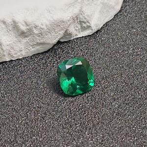 May include: A faceted, emerald-green gemstone with a cushion cut. The gem is set against a dark, textured surface, with a white, rough-textured element in the background. The gem's facets reflect light, creating a brilliant sparkle.
