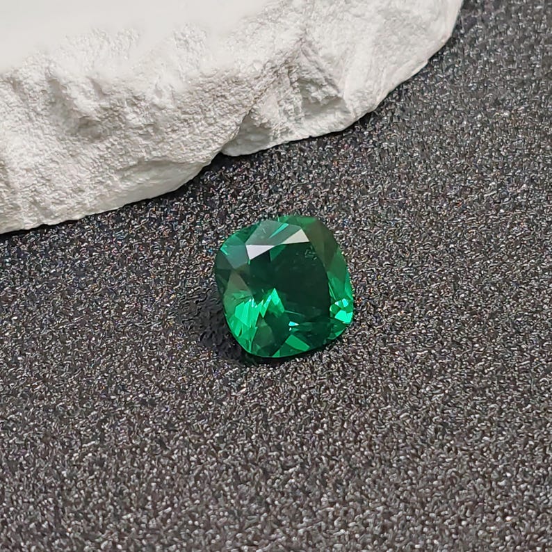 May include: A faceted, emerald-green gemstone with a cushion cut. The gem is set against a dark, textured surface, with a white, rough-textured element in the background. The gem's facets reflect light, creating a brilliant sparkle.