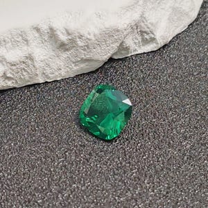 May include: A faceted, emerald-green gemstone with a cushion cut. The gem is displayed on a dark, textured surface, with a white, rough-textured element in the background. The gem's facets reflect light, creating a sparkling effect.