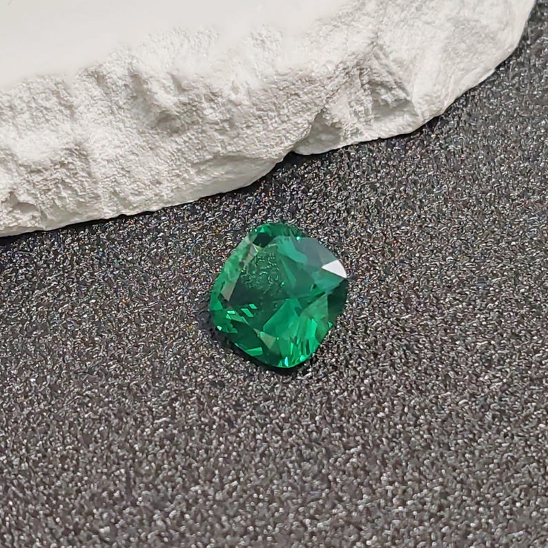 May include: A faceted, emerald-green gemstone with a cushion cut. The gem is displayed on a dark, textured surface, with a white, rough-textured element in the background. The gem's facets reflect light, creating a sparkling effect.