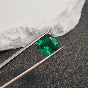 May include: A vibrant, emerald-green gemstone, held by silver tweezers. The cushion-cut gem displays facets that reflect light, set against a textured white surface and a dark background. A close-up shot highlighting the gem's clarity and color.