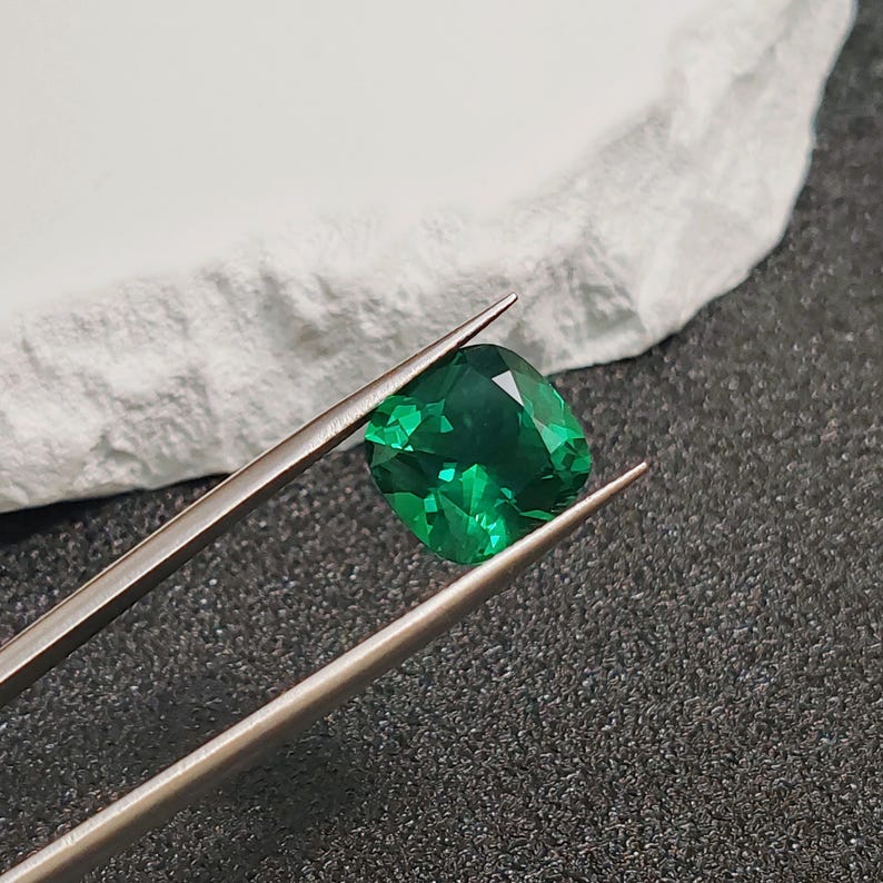 May include: A vibrant, emerald-green gemstone, held by silver tweezers. The cushion-cut gem displays facets that reflect light, set against a textured white surface and a dark background. A close-up shot highlighting the gem's clarity and color.