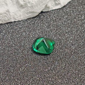 May include: A faceted, emerald-green gemstone with a cushion cut. The gem is displayed on a textured, dark gray surface, with a white fabric in the background. The gemstone's facets reflect light, creating a sparkling effect.