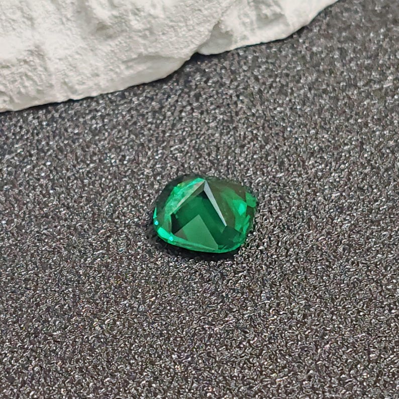 May include: A faceted, emerald-green gemstone with a cushion cut. The gem is displayed on a textured, dark gray surface, with a white fabric in the background. The gemstone's facets reflect light, creating a sparkling effect.