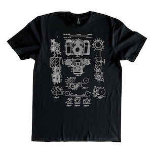 Puede incluir: Camiseta negra con un estampado blanco de un diseño de patente de cámara. El número de patente es 5.604.411 y la fecha es 13 de mayo de 1997.