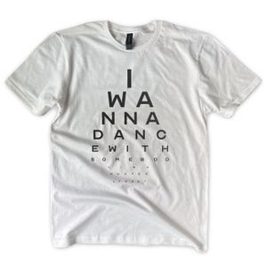 May include: White t-shirt with the text "I WANNA DANCE WITH SOMEBOD..." in a black, eye chart style. The shirt has short sleeves and a crew neck. The text is centered on the front of the shirt.