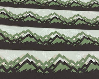 May include: Four layered paper cutouts of mountain ranges in shades of brown, green, and white. The paper cutouts are arranged horizontally, creating a repeating pattern of peaks and valleys. The background is a light, floral-patterned paper.