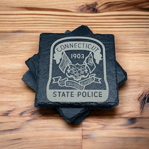 May include: Four black slate coasters with a white engraved design of the Connecticut State Police emblem. The emblem features the state seal, the words "Connecticut" and "State Police", and the year "1903".