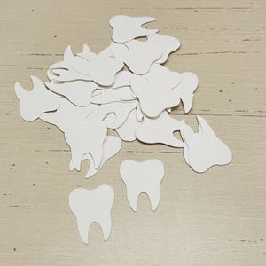 May include: A collection of white paper teeth cutouts scattered on a light wood-grain surface. The teeth vary in size and shape, with some resembling molars and others incisors. The dental-themed cutouts are arranged in a random pattern.