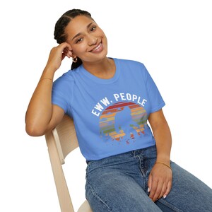 May include: A woman wearing a light blue t-shirt with a retro sunset graphic and the text "EWW, PEOPLE".