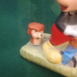 Op de afbeelding: Een keramieken beeldje met Mickey Mouse in een klassieke pose, met een rode broek, gele schoenen en zwarte handschoenen. Er is ook een klein figuurtje van een aap aanwezig. De basis is lichtgroen.
