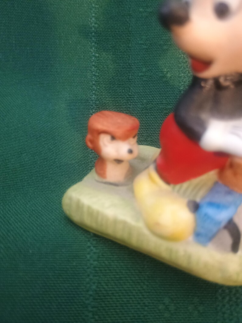 Op de afbeelding: Een keramieken beeldje met Mickey Mouse in een klassieke pose, met een rode broek, gele schoenen en zwarte handschoenen. Er is ook een klein figuurtje van een aap aanwezig. De basis is lichtgroen.