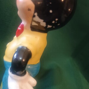 May include: A vintage figurine of a cartoon character, featuring a black head with white patches, a yellow shirt, blue pants, and a red bow tie. The figurine's arm is black with a white glove. The paint shows signs of wear.