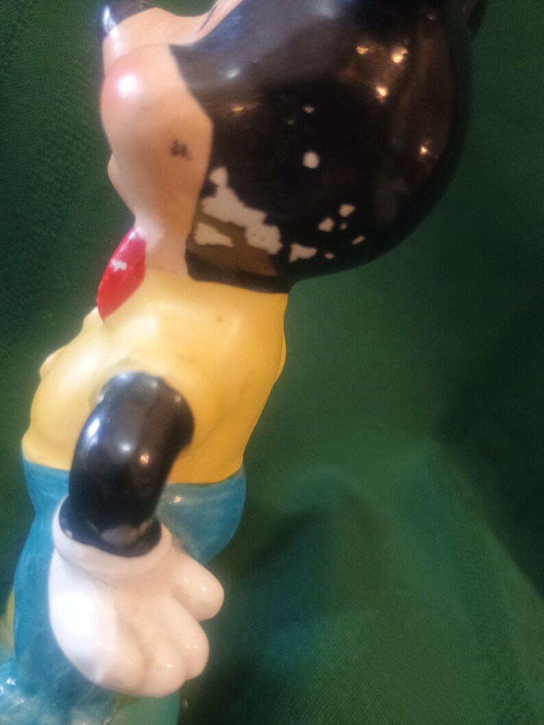 May include: A vintage figurine of a cartoon character, featuring a black head with white patches, a yellow shirt, blue pants, and a red bow tie. The figurine's arm is black with a white glove. The paint shows signs of wear.