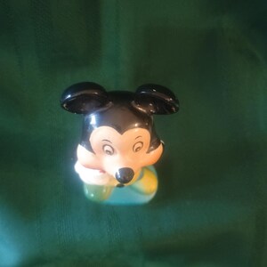 May include: A ceramic Mickey Mouse figurine. The figurine has black ears, a black nose, and a cream-colored face. Mickey is wearing a blue and yellow outfit and holding a green object. The figurine is set against a green background.