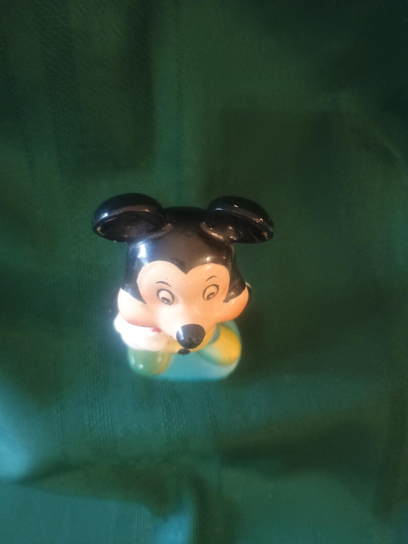 May include: A ceramic Mickey Mouse figurine. The figurine has black ears, a black nose, and a cream-colored face. Mickey is wearing a blue and yellow outfit and holding a green object. The figurine is set against a green background.