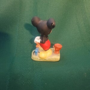 Op de afbeelding: Een klein beeldje van Mickey Mouse, van achteren gezien. Mickey draagt een rode broek, gele schoenen en witte handschoenen. Het beeldje staat op een zanderige basis met kleine, kleurrijke accessoires.