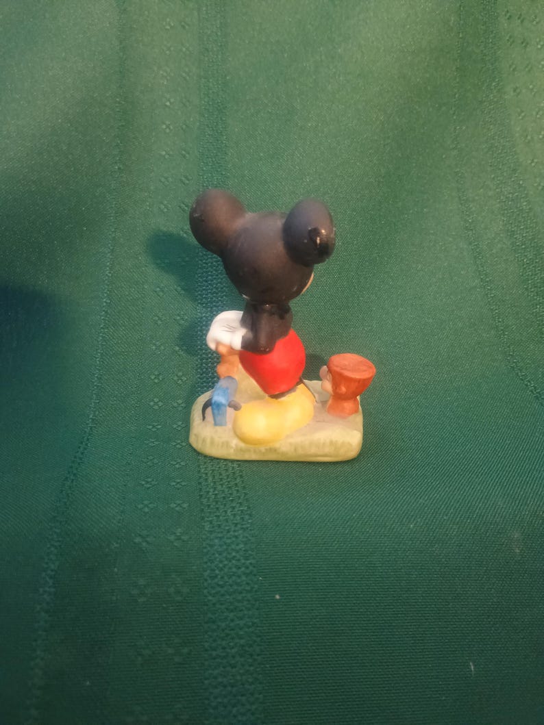 Op de afbeelding: Een klein beeldje van Mickey Mouse, van achteren gezien. Mickey draagt een rode broek, gele schoenen en witte handschoenen. Het beeldje staat op een zanderige basis met kleine, kleurrijke accessoires.