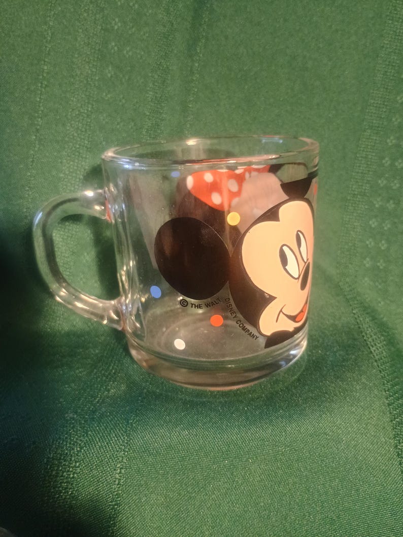Pu&ograve; includere: Tazza di vetro trasparente con Topolino. La tazza ha un manico e un design colorato del volto di Topolino e pois. Le parole "The Walt Disney Company" sono stampate sulla tazza.