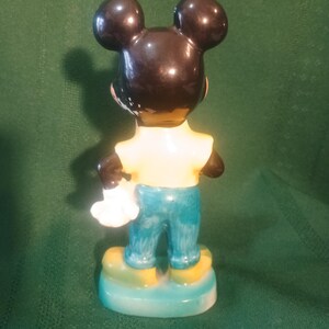 May include: A vintage Mickey Mouse figurine, viewed from the back. The figure has black ears and head, a yellow shirt, blue pants, and yellow shoes. The figurine is holding a white glove and stands on a blue and yellow base.