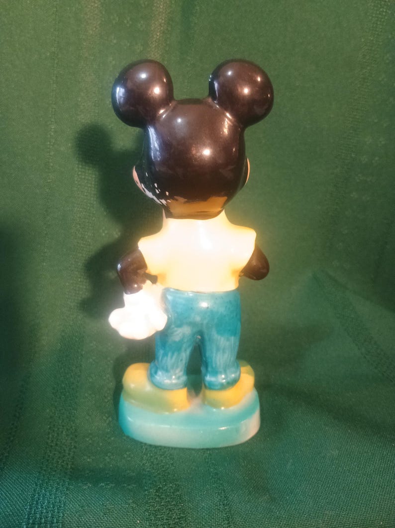 May include: A vintage Mickey Mouse figurine, viewed from the back. The figure has black ears and head, a yellow shirt, blue pants, and yellow shoes. The figurine is holding a white glove and stands on a blue and yellow base.