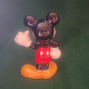 Puede incluir: Una peque&ntilde;a figura de Mickey Mouse. La figura tiene cabeza y orejas negras, una chaqueta roja con dos botones blancos, guantes blancos y zapatos amarillos. La figura est&aacute; posando con el pulgar hacia arriba.