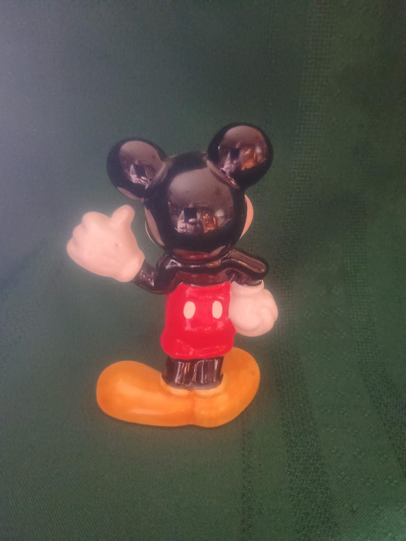 Puede incluir: Una peque&ntilde;a figura de Mickey Mouse. La figura tiene cabeza y orejas negras, una chaqueta roja con dos botones blancos, guantes blancos y zapatos amarillos. La figura est&aacute; posando con el pulgar hacia arriba.