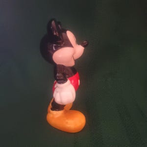 Puede incluir: Figura de cer&aacute;mica de Mickey Mouse. Tiene orejas y cabeza negras, cara blanca, pantalones cortos rojos, guantes blancos y zapatos amarillos. La figura se ve de lado, con una ligera inclinaci&oacute;n de la cabeza. El fondo es verde oscuro.