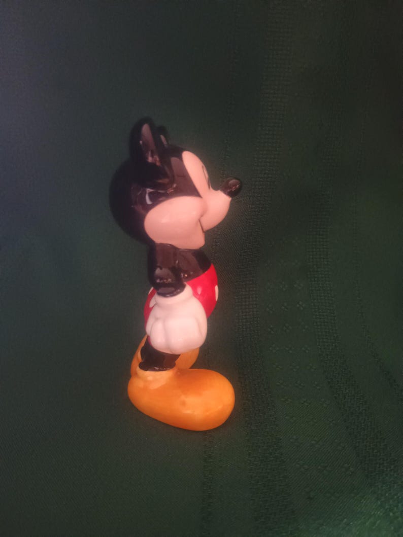 Puede incluir: Figura de cer&aacute;mica de Mickey Mouse. Tiene orejas y cabeza negras, cara blanca, pantalones cortos rojos, guantes blancos y zapatos amarillos. La figura se ve de lado, con una ligera inclinaci&oacute;n de la cabeza. El fondo es verde oscuro.