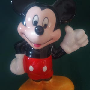 Puede incluir: Una figura de cer&aacute;mica de Mickey Mouse. La figura tiene orejas negras, cuerpo negro, cara rosa, guantes blancos, pantalones cortos rojos con dos botones blancos y zapatos amarillos. Mickey est&aacute; haciendo una se&ntilde;al de aprobaci&oacute;n.