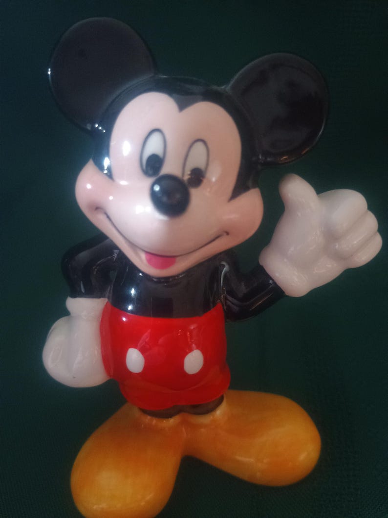 Puede incluir: Una figura de cer&aacute;mica de Mickey Mouse. La figura tiene orejas negras, cuerpo negro, cara rosa, guantes blancos, pantalones cortos rojos con dos botones blancos y zapatos amarillos. Mickey est&aacute; haciendo una se&ntilde;al de aprobaci&oacute;n.
