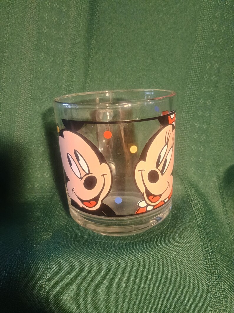 Pu&ograve; includere: Bicchiere di vetro trasparente con i personaggi dei cartoni animati di Topolino e Minnie. Il bicchiere ha un bordo nero e pois colorati. I personaggi dei cartoni animati sono rosa, neri, rossi e bianchi.