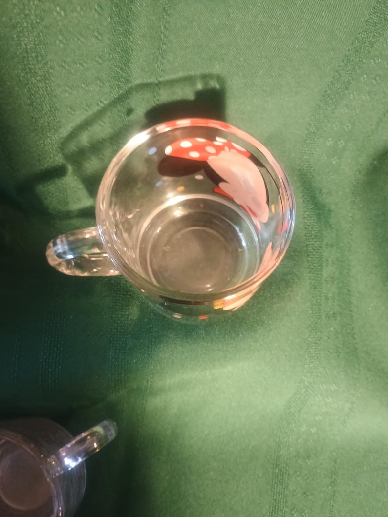 Pu&ograve; includere: Tazza di vetro trasparente con manico, con un disegno di fungo dipinto con pois rossi e bianchi. La tazza &egrave; vuota e poggia su una superficie verde.
