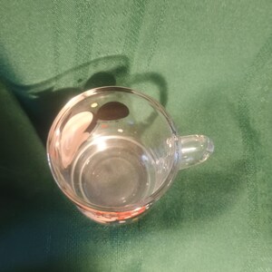 Pu&ograve; includere: Una tazza di vetro trasparente con un piccolo manico. La tazza &egrave; vuota e si trova su una superficie verde. Il fondo della tazza ha un design rosso e arancione.