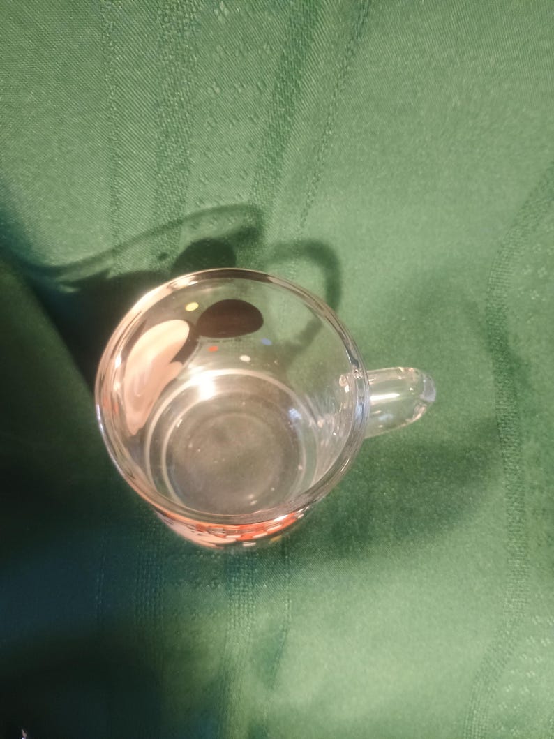 Pu&ograve; includere: Una tazza di vetro trasparente con un piccolo manico. La tazza &egrave; vuota e si trova su una superficie verde. Il fondo della tazza ha un design rosso e arancione.