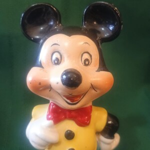 May include: A vintage Mickey Mouse figurine. The figure has black ears, a black nose, and a smiling face with rosy cheeks. Mickey wears a yellow shirt with black buttons, a red bow tie, white gloves, and blue pants. The figurine is a collectible item.