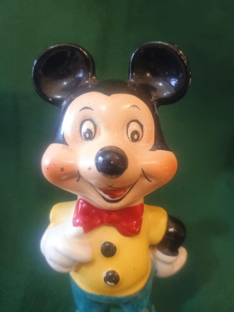 May include: A vintage Mickey Mouse figurine. The figure has black ears, a black nose, and a smiling face with rosy cheeks. Mickey wears a yellow shirt with black buttons, a red bow tie, white gloves, and blue pants. The figurine is a collectible item.
