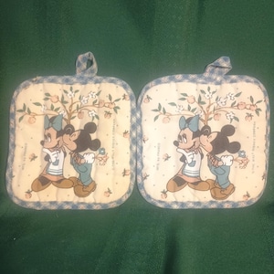 May include: Two square pot holders featuring Mickey and Minnie Mouse embracing under a tree with fruit. The pot holders have a light blue and white gingham border and a loop for hanging.