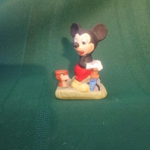 Op de afbeelding: Een keramieken beeldje van Mickey Mouse in een klassieke pose, gekleed in een rode broek, gele schoenen en witte handschoenen. Het beeldje bevat een klein beeldje van Donald Duck en een blauw object. De basis is lichtbruin.
