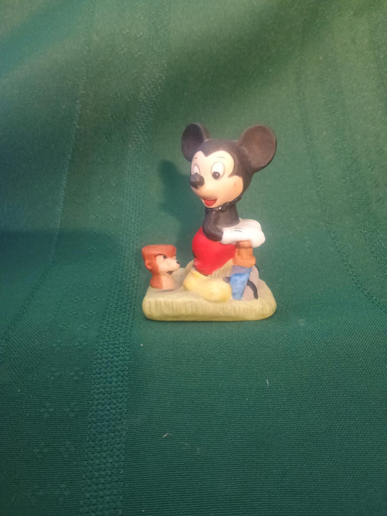 Op de afbeelding: Een keramieken beeldje van Mickey Mouse in een klassieke pose, gekleed in een rode broek, gele schoenen en witte handschoenen. Het beeldje bevat een klein beeldje van Donald Duck en een blauw object. De basis is lichtbruin.