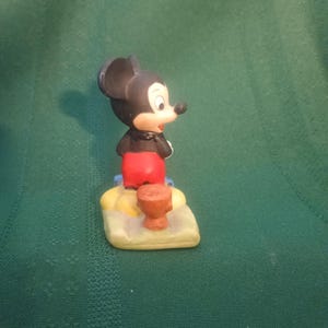 Op de afbeelding: Een keramieken Mickey Mouse beeldje, met zwarte oren, rode broek en witte handschoenen. Hij staat op een gele en beige basis met een klein bruin object. Een verzamelobject.
