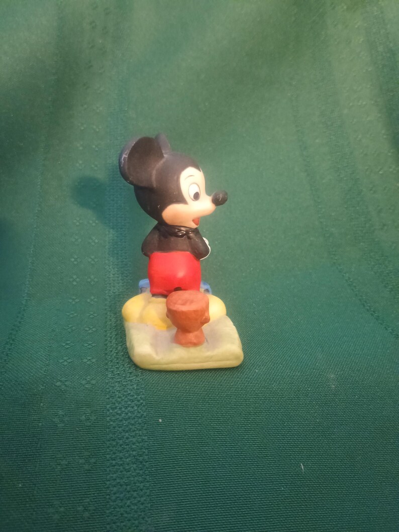 Op de afbeelding: Een keramieken Mickey Mouse beeldje, met zwarte oren, rode broek en witte handschoenen. Hij staat op een gele en beige basis met een klein bruin object. Een verzamelobject.