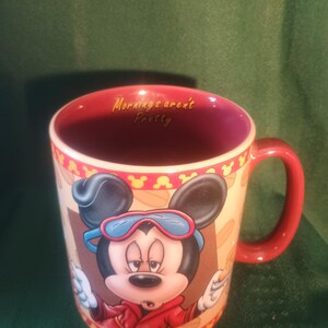 Może przedstawiać: Ceramiczny kubek do kawy z czerwonym uchwytem i wnętrzem, przedstawiający Myszka Miki w czerwonej kurtce i okularach. Kubek ma napis "Mornings aren't Pretty" w środku. Zewnętrzna strona ma beżowe tło z powtarzającym się wzorem.