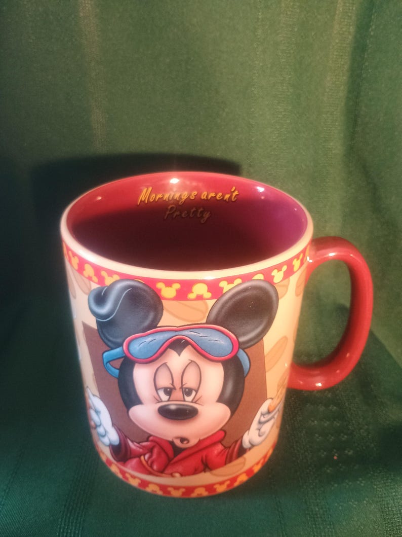 Może przedstawiać: Ceramiczny kubek do kawy z czerwonym uchwytem i wnętrzem, przedstawiający Myszka Miki w czerwonej kurtce i okularach. Kubek ma napis "Mornings aren't Pretty" w środku. Zewnętrzna strona ma beżowe tło z powtarzającym się wzorem.