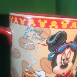 Może przedstawiać: Ceramiczny kubek do kawy z Myszka Miki w czerwonym szlafroku i niebieskiej masce do spania. Kubek ma napis "Morning Ear Droop" i "Cheesy Robe". Obręcz jest czerwona z ż&oacute;łtymi sylwetkami Myszki Miki.