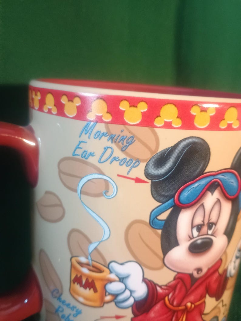 Może przedstawiać: Ceramiczny kubek do kawy z Myszka Miki w czerwonym szlafroku i niebieskiej masce do spania. Kubek ma napis "Morning Ear Droop" i "Cheesy Robe". Obręcz jest czerwona z ż&oacute;łtymi sylwetkami Myszki Miki.