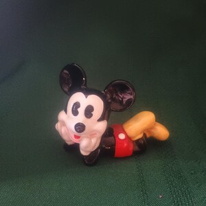 May include: A small, glossy figurine of Mickey Mouse in a relaxed pose. The figure features black ears, a white face, and a red shorts. The figure is lying on its stomach with yellow shoes and white gloves.
